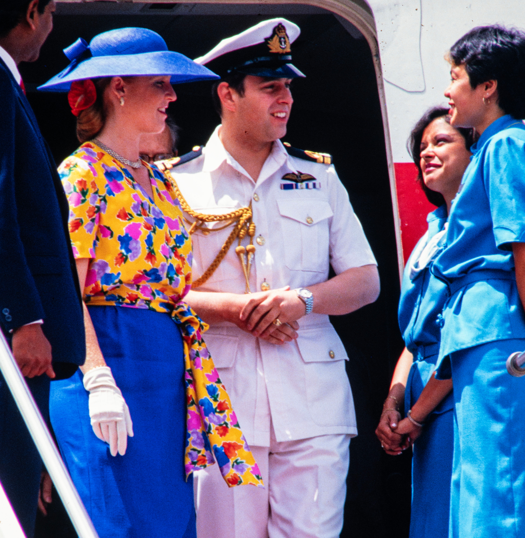 Sarah Ferguson wearing a blue skirt and hat and yellow floral top standing on the stairs of a plane next to Prince Andrew in a naval uniform 