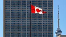 A Canadian flag, flying on a tall pole in front of a skyscraper.