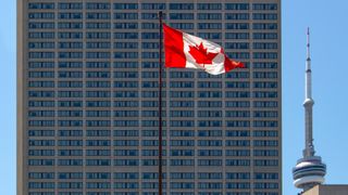 A Canadian flag, flying on a tall pole in front of a skyscraper.