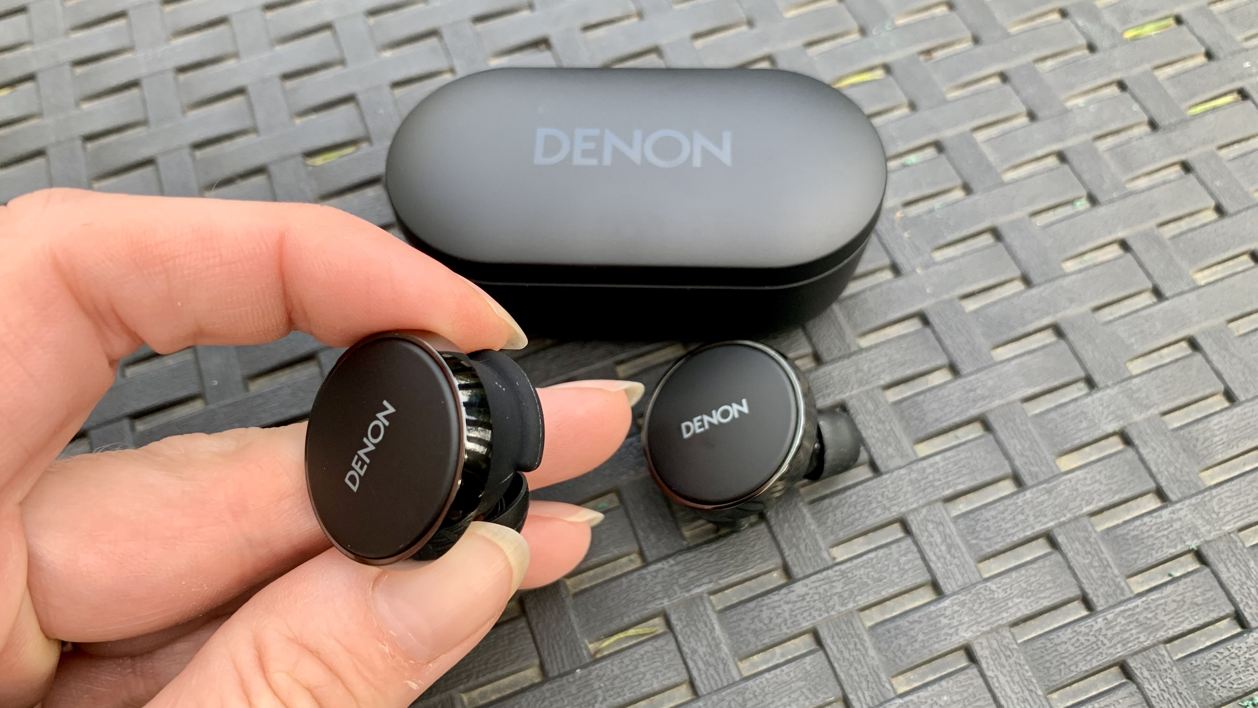 Denon PerL Pro earbud held in a hand, with the case in the background on a black table