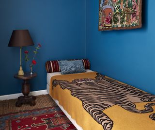 A bedroom painted in Benjamin Moore's bright blue Deep Ocean shade which beautifully contrasts with the yellow tiger bedding, red rug and multicolor artwork on the wall; there is also a dark wood bedside table with a table lamp and flowers in a vase