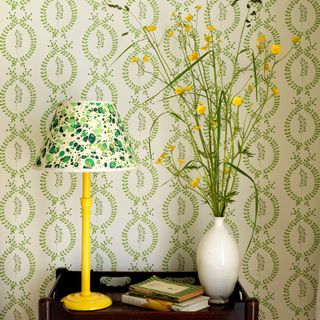 A yellow table lamp with a marbled green lampshade next to a vase of flowers with a botanical-print wallpaper as a backdrop