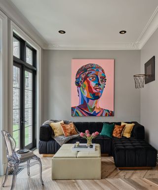 A living room with a black sectional, a beige ottoman, and a bright pink piece of art on the wall