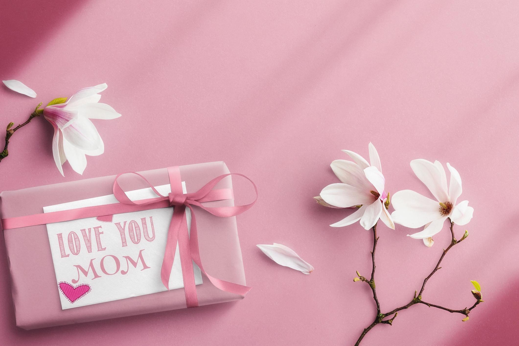  a pink gift parcel that says I love you mom, next to flowers on a pink background  
