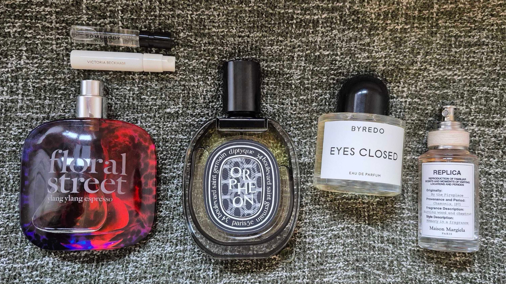 A selection of some of the winter perfumes featured in this guide, including two testers of PHLUR&#039;s Somebody Wood and Victoria Beckham Beauty&#039;s Suite 302 fragrances and full sizes of Floral Streets Ylang Ylang Espresso, Diptyque Orpheon, Byredo Eyes Closed and Maison Margiela&#039;s By The Fireplace perfumes, all spread in a line across a green marl blanket background.