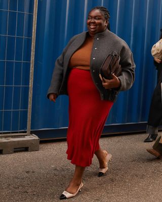 Woman wears a red midi skirt, tan top, grey bomber jacket and slingback kitten heels.