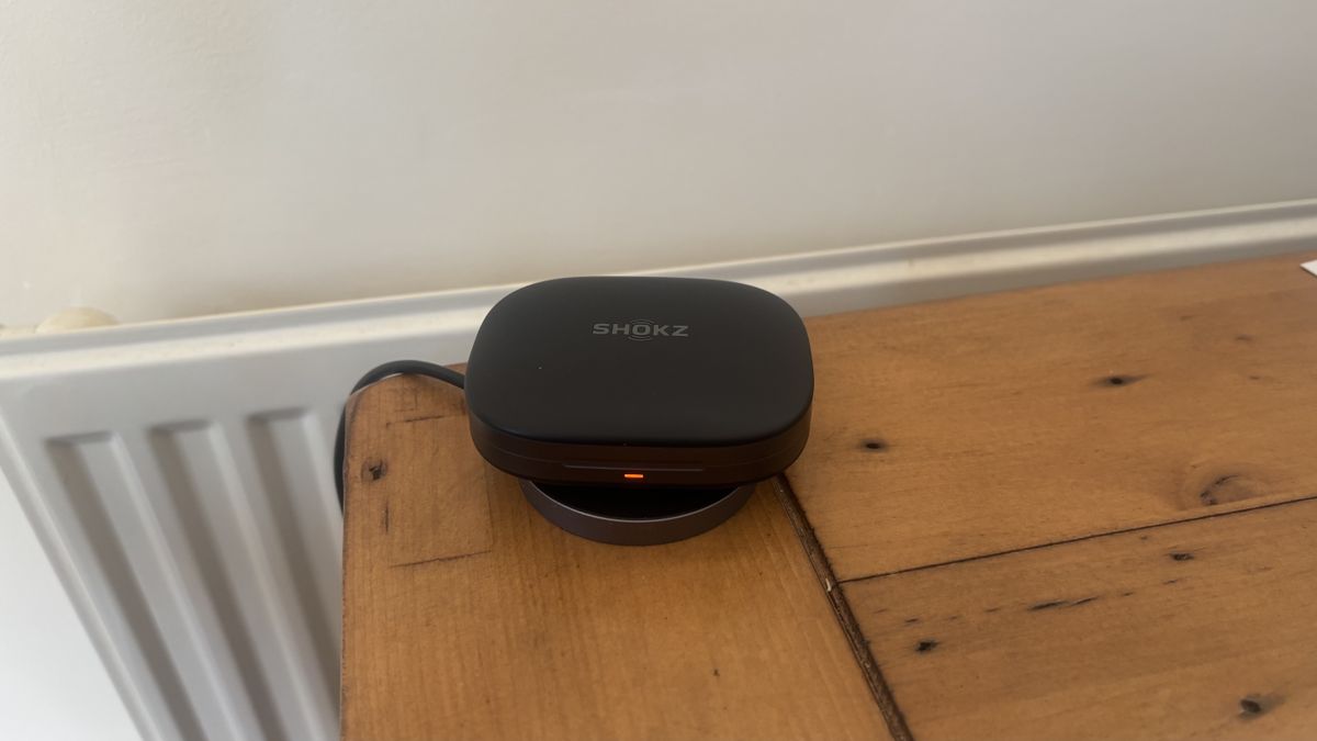 Shokz OpenFit Pro open earbuds charging case, using a wireless charger, on a wooden surface.