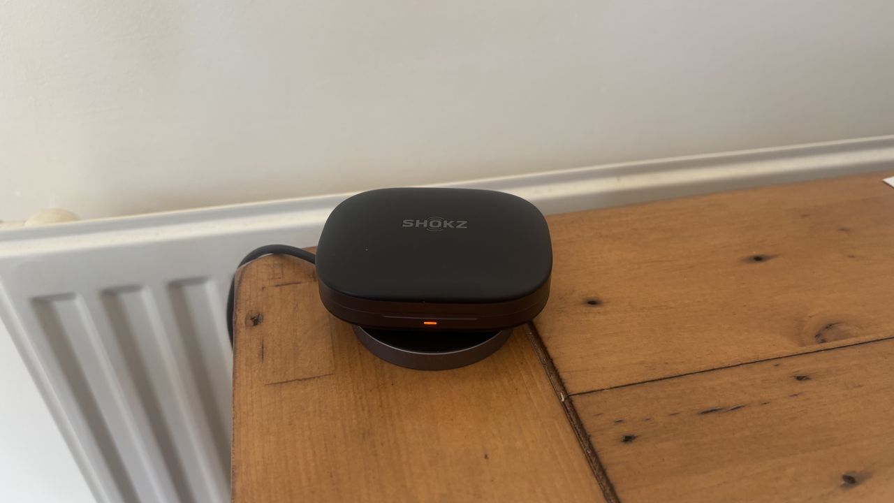Shokz OpenFit Pro open earbuds charging case, using a wireless charger, on a wooden surface.