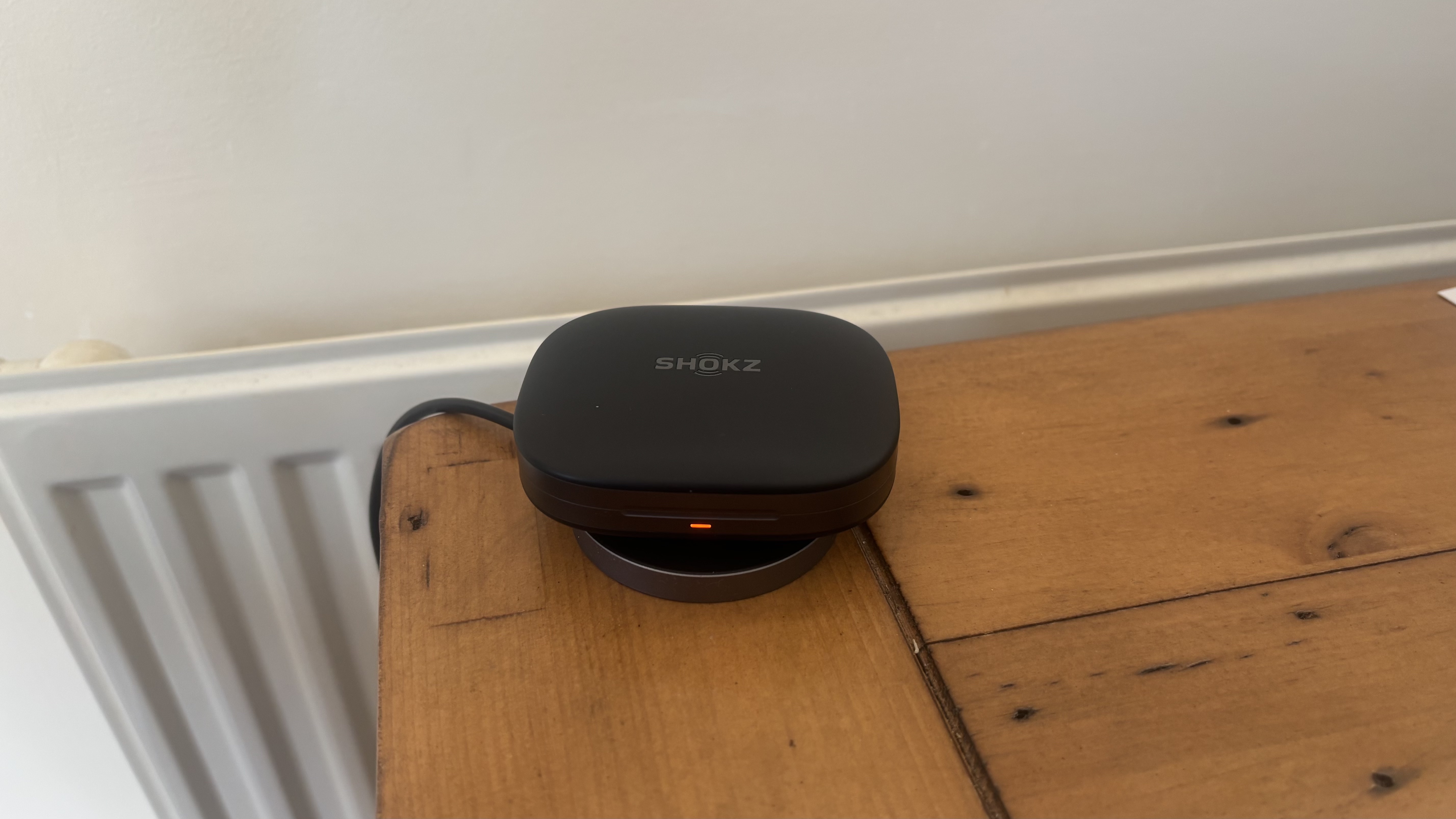 Shokz OpenFit Pro open earbuds charging case, using a wireless charger, on a wooden surface.