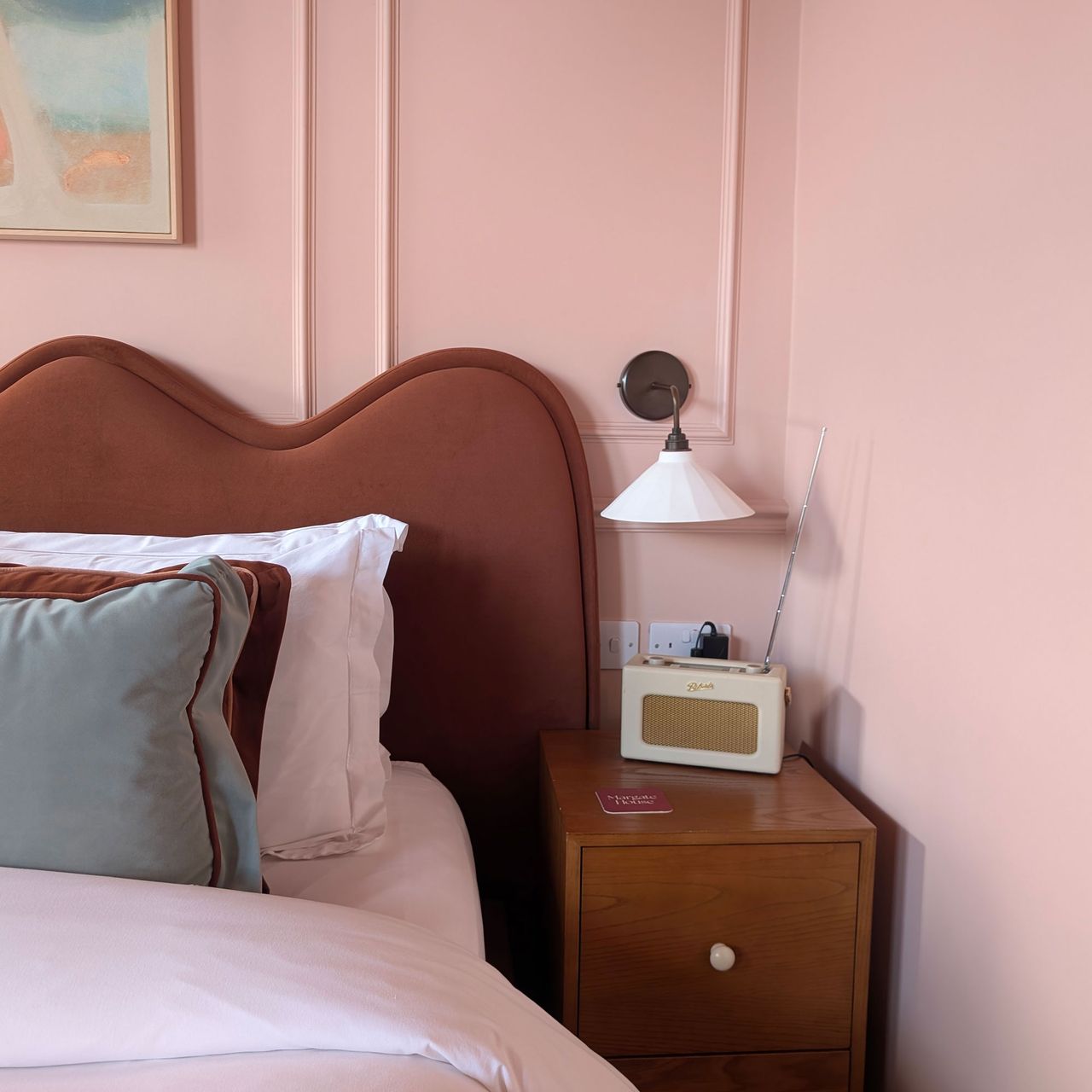 Close up of brown wavy bed headboard beside white shaded wall light above cream Roberts Radio on wooden bedside table