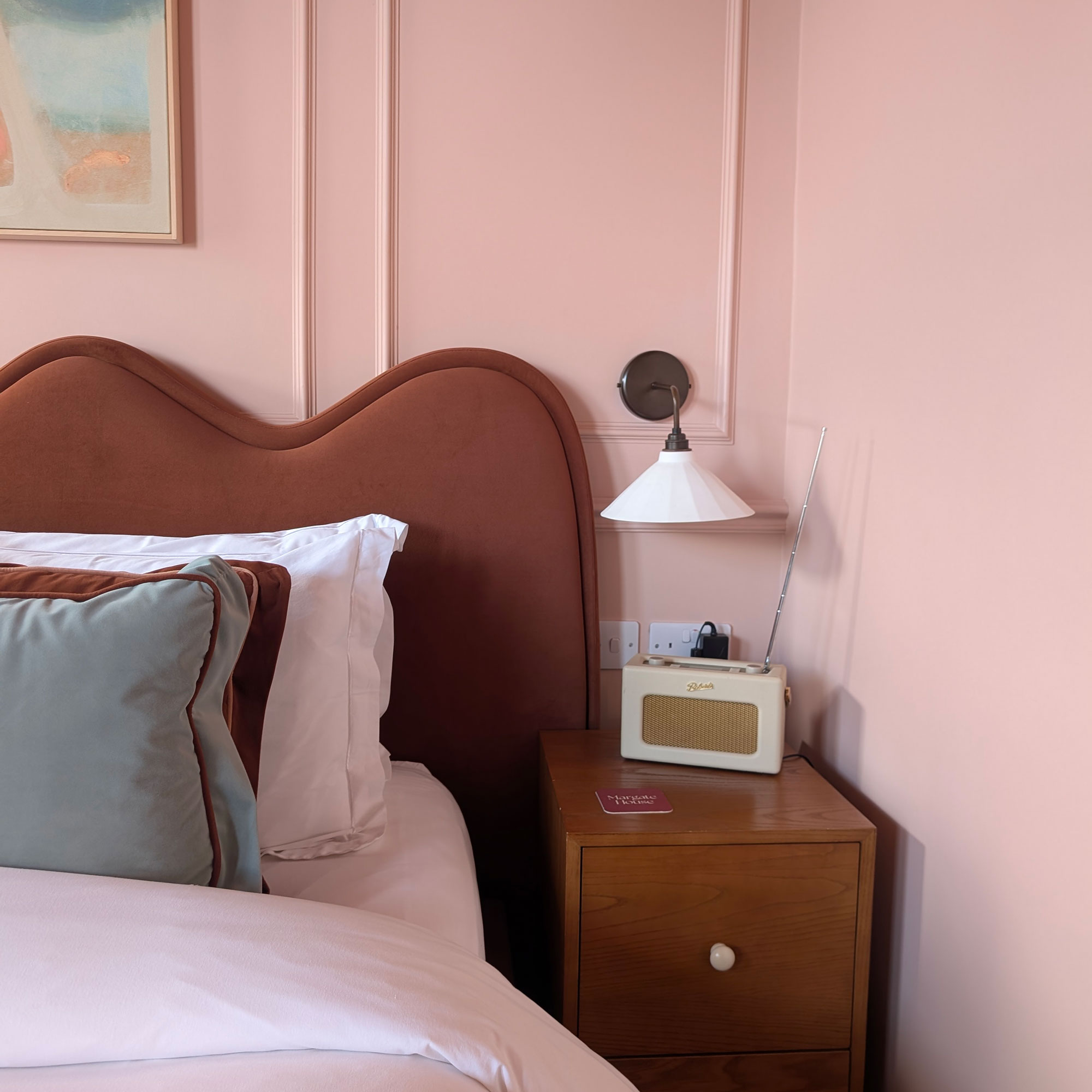 Close up of brown wavy bed headboard beside white shaded wall light above cream Roberts Radio on wooden bedside table