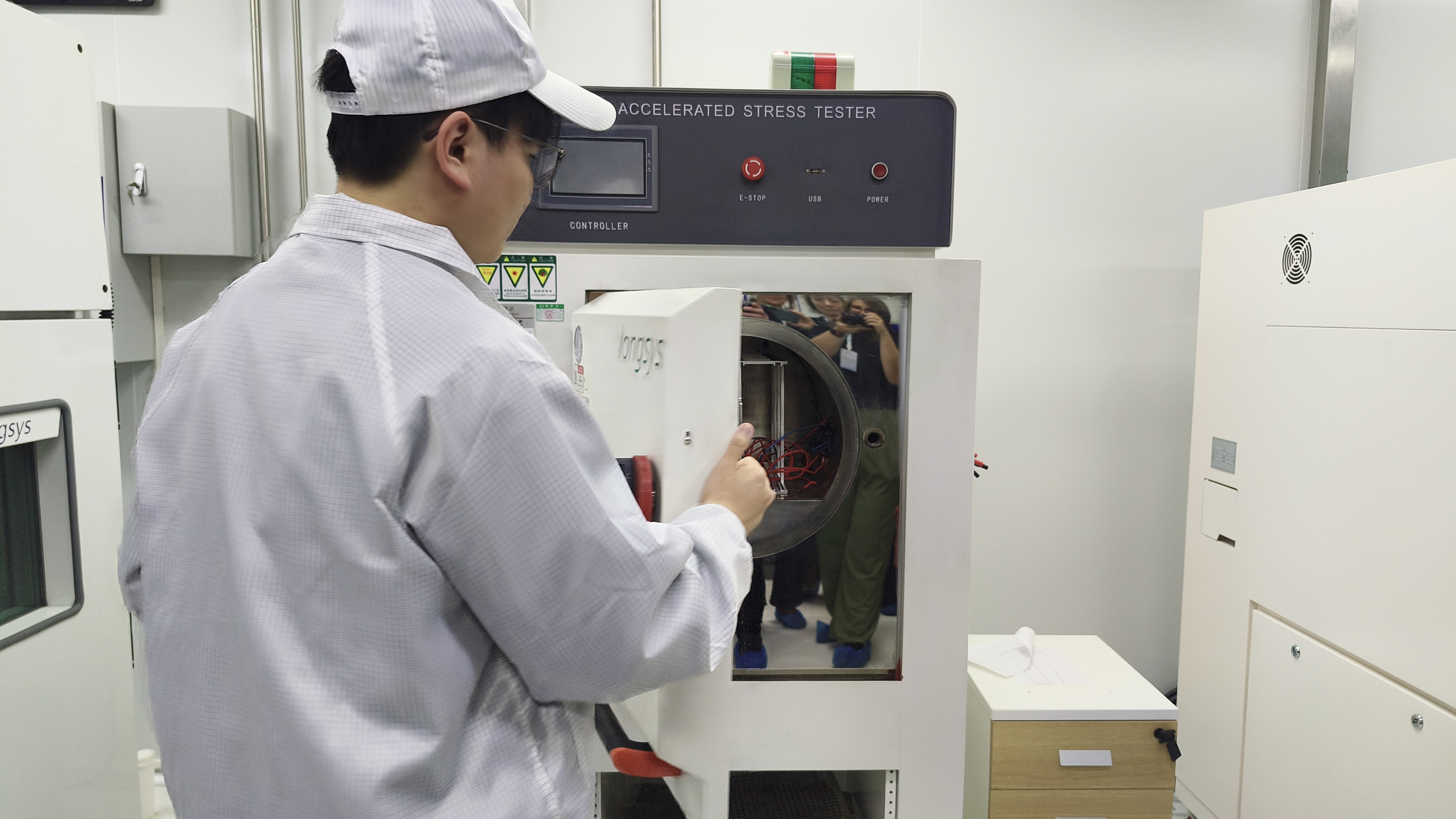 A laboratory worker in a white uniform opens the circular door of a machine labeled "Highly Accelerated Stress Tester."
