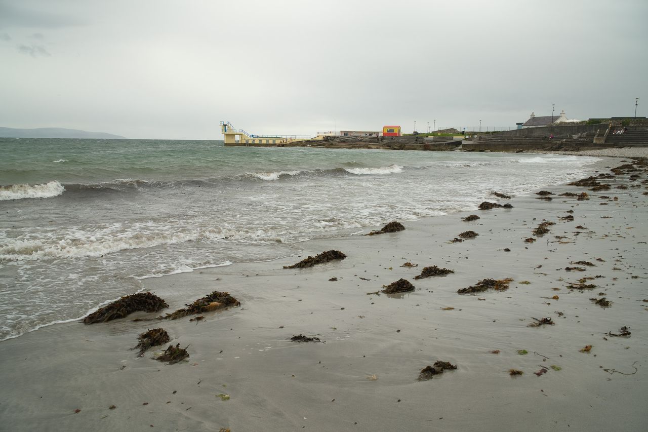 Seaweed is scattered across a wet beach as waves roll toward a concrete seafront and diving platform.
