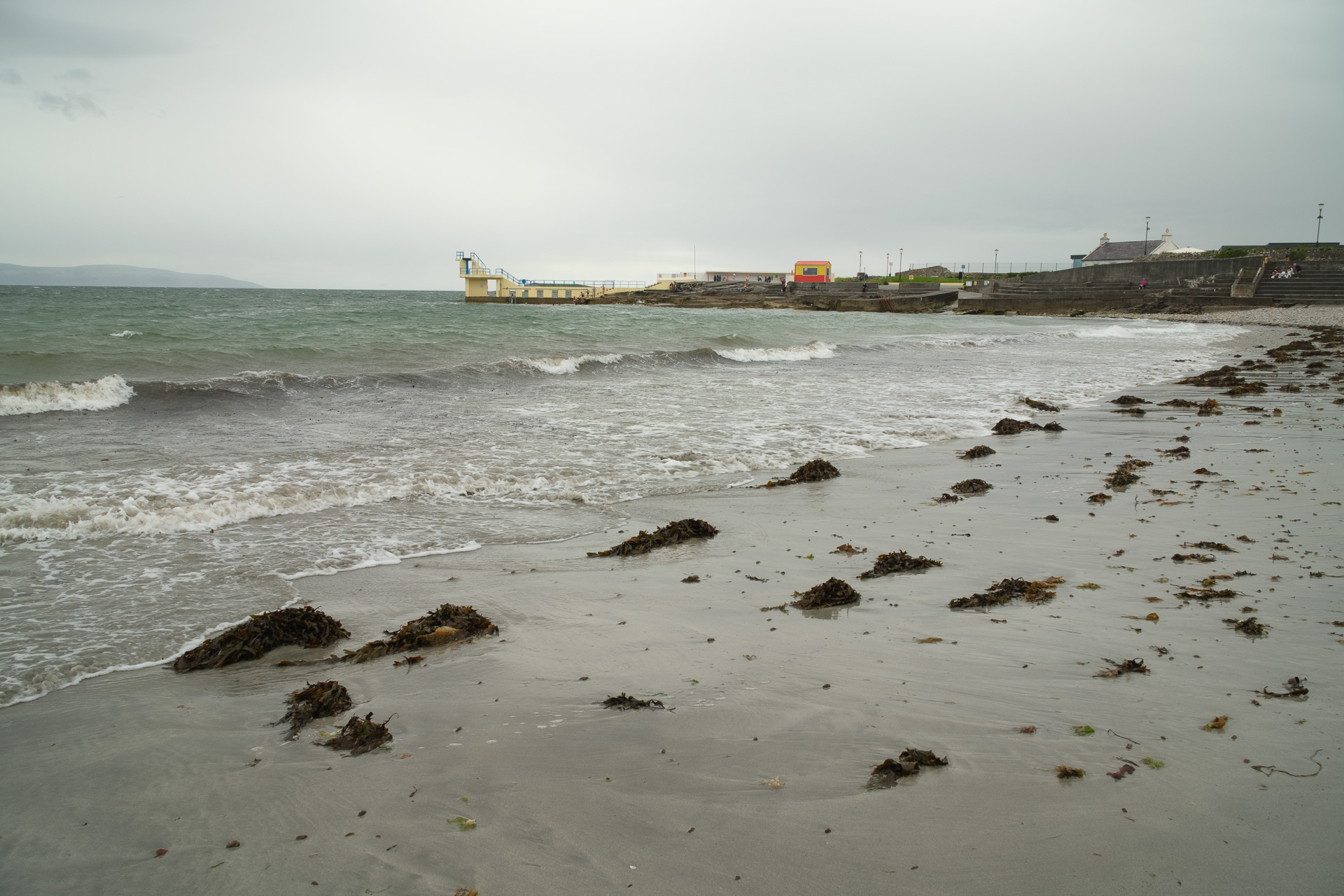 Seaweed is scattered across a wet beach as waves roll toward a concrete seafront and diving platform.