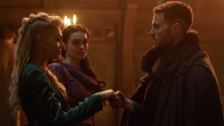 Guinevere (Jordan Alexandra) in a green dress and Ceinwyn (Emily John) in a red dress greet Arthur (Iain De Caestecker) in a fur cloak in The Winter King.