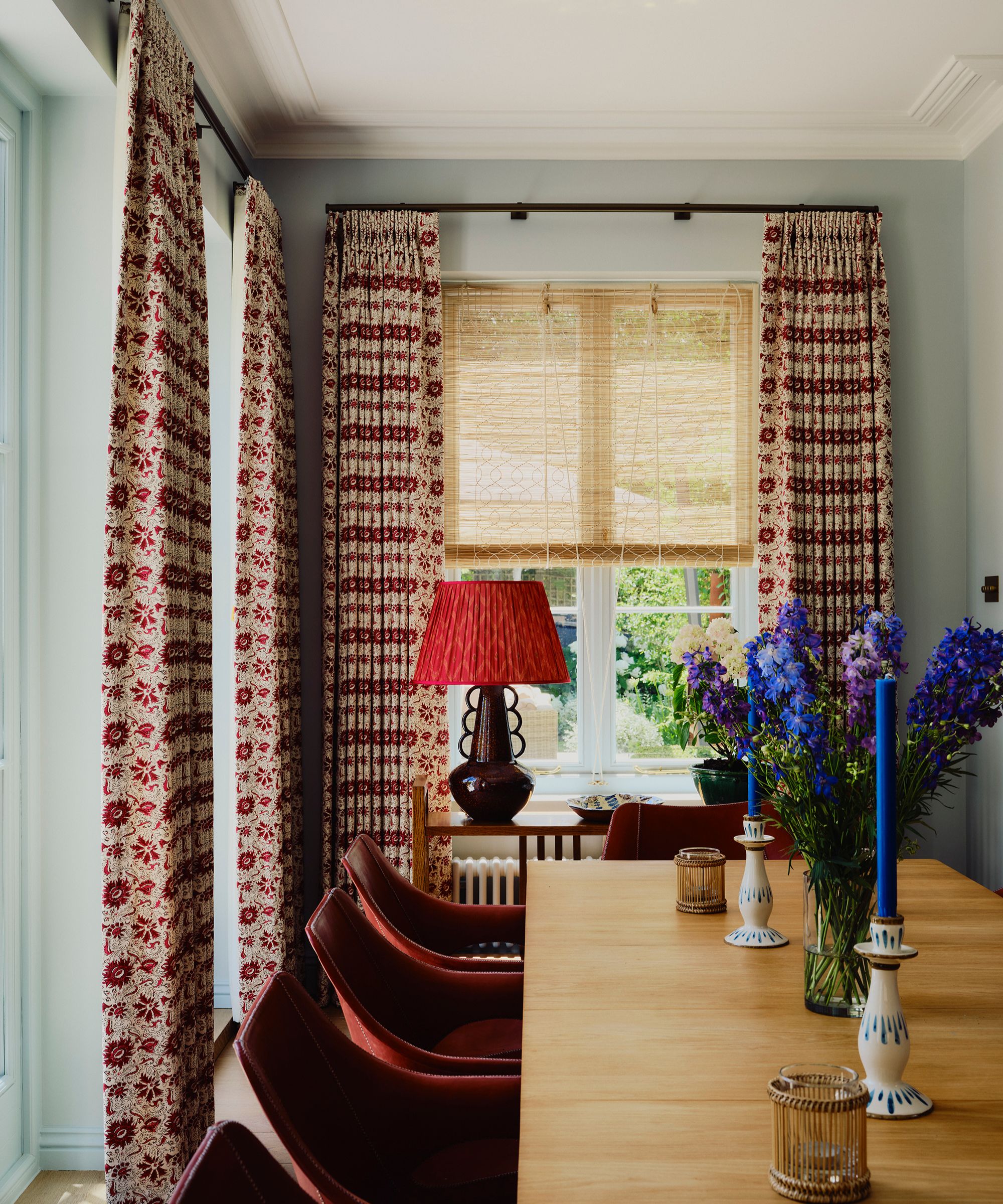 Dining room with pale blue walls, a window with a rattan blind, and red floral drapes on top, a wooden dining table and red dining chairs underneath, decorated with blue and white candlesticks