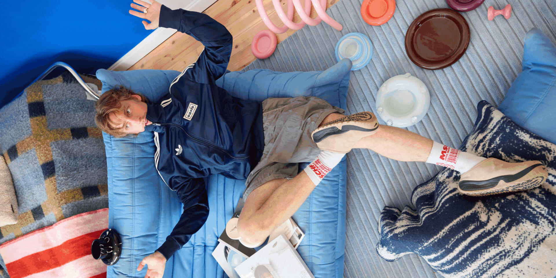 A rotating gif depicting, in order of appearance, a young man dressed in sporty-chic clothing, sitting on a pale blue retro sofa, colorful, plump, sheeny objects in different pastel shades, a filled-with-collectibles bedroom, and more eye-catching, pink items showcased next to a plate filled with fuzzy, flat peaches.