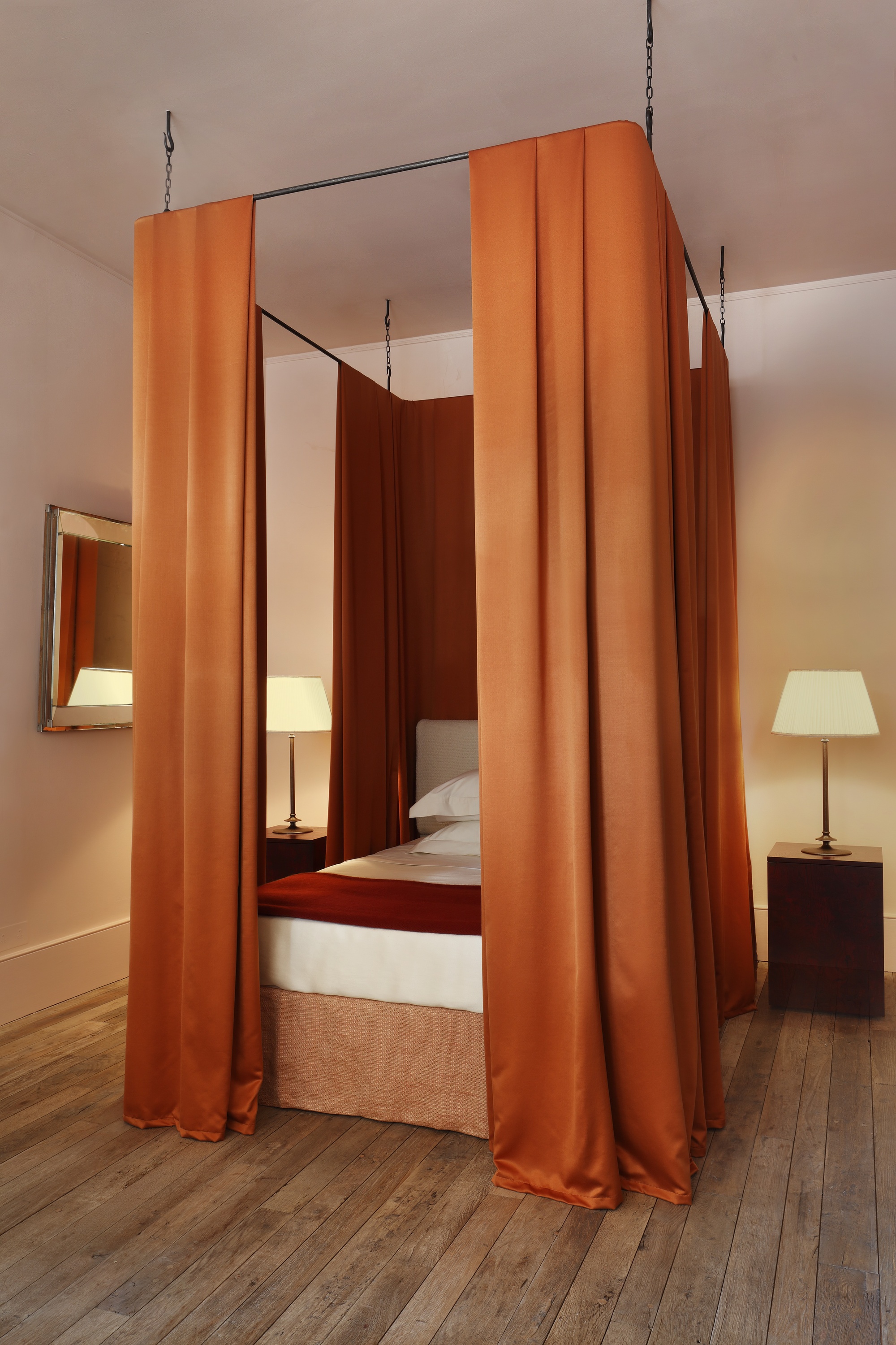 modern bedroom with canopy bed with orange curtains, timber floors, timber bedside tables with lamps, and a mirror on the wall