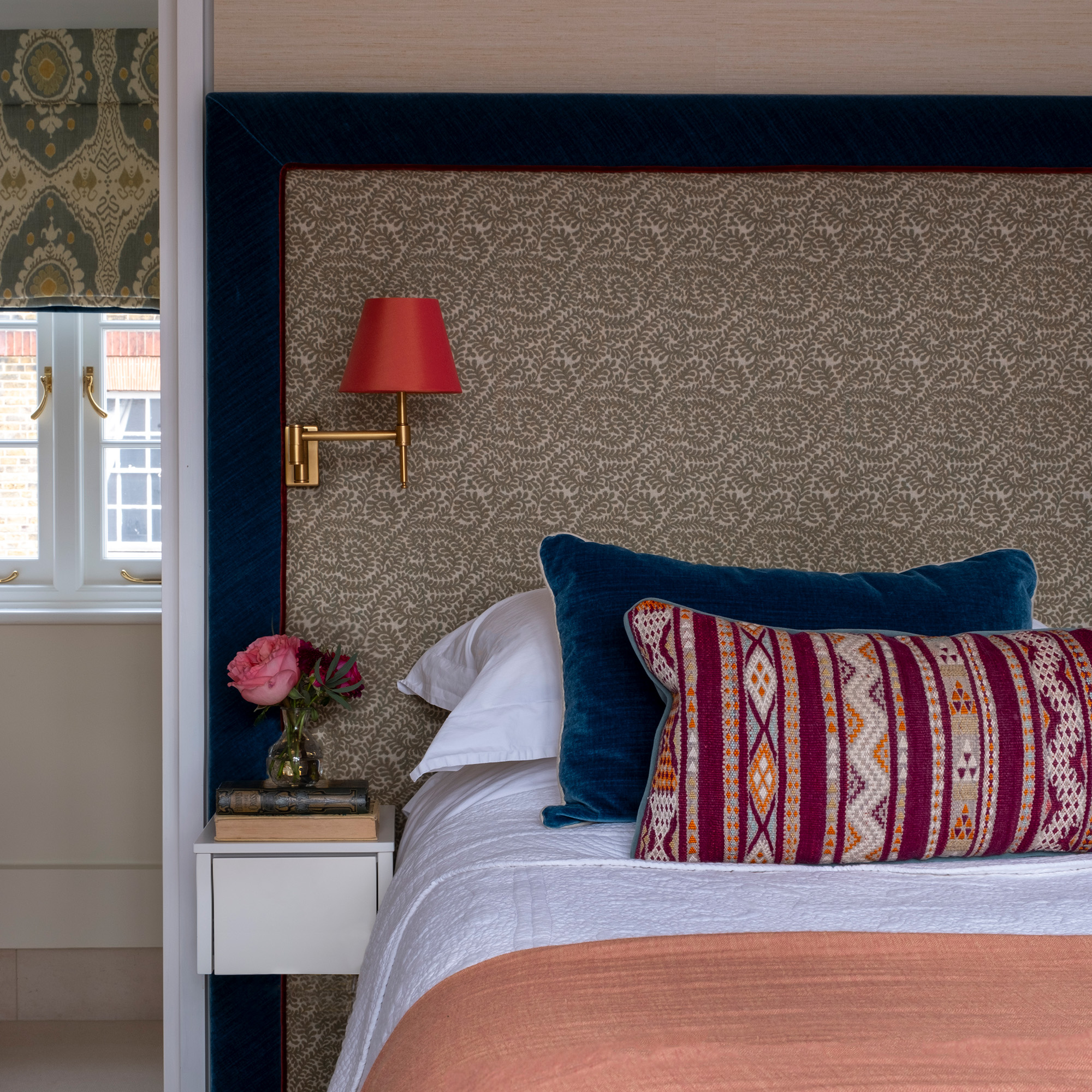bedroom with patterned oversized headboard with pale grey pattern and velvet blue border attached to the headboard is a brass wall light with orange shade