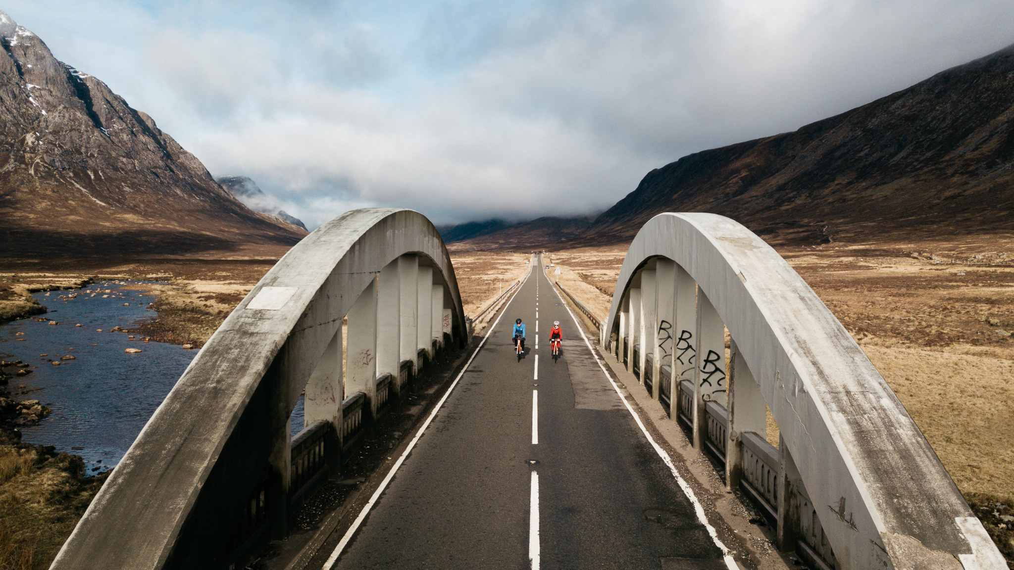 Two cyclists captured by a drone riding over a bridge in Glencoe, Scotland