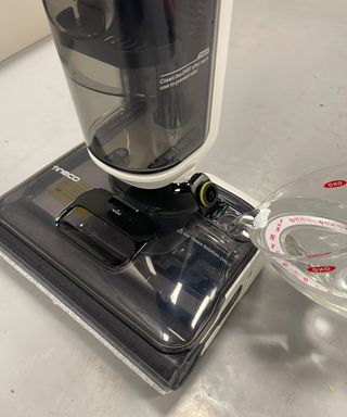 Cleaning solution being poured out of clear jug into Tineco Floor One Stretch S6's clean water tank in the floorhead, on gray floor in Future's test center