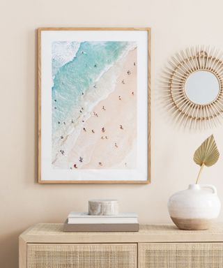 A coastal wall art print with a birds eye sea and sand scene with a wooden frame, a starburst wooden mirror next to it, and a rattan sideboard with two books and a ceramic vase with a leaf in it on top
