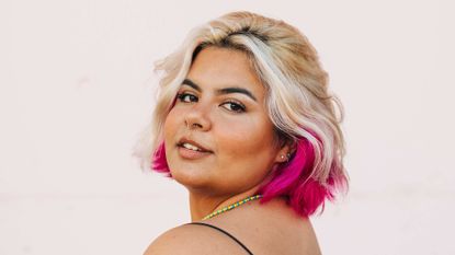 A woman is seen standing against the wall, she had chemically dyes hair in blonde and pink