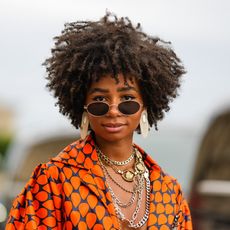 Sarah Monteil wears black sunglasses, oversized silver earrings, silver large chain necklaces, a navy blue with orange print pattern oversized silk shirt, high waist matching navy blue with orange print pattern large pants, outside the Thom Browne show, during Paris Fashion Week - Menswear Spring/Summer 2023, on June 26, 2022 in Paris, France. (