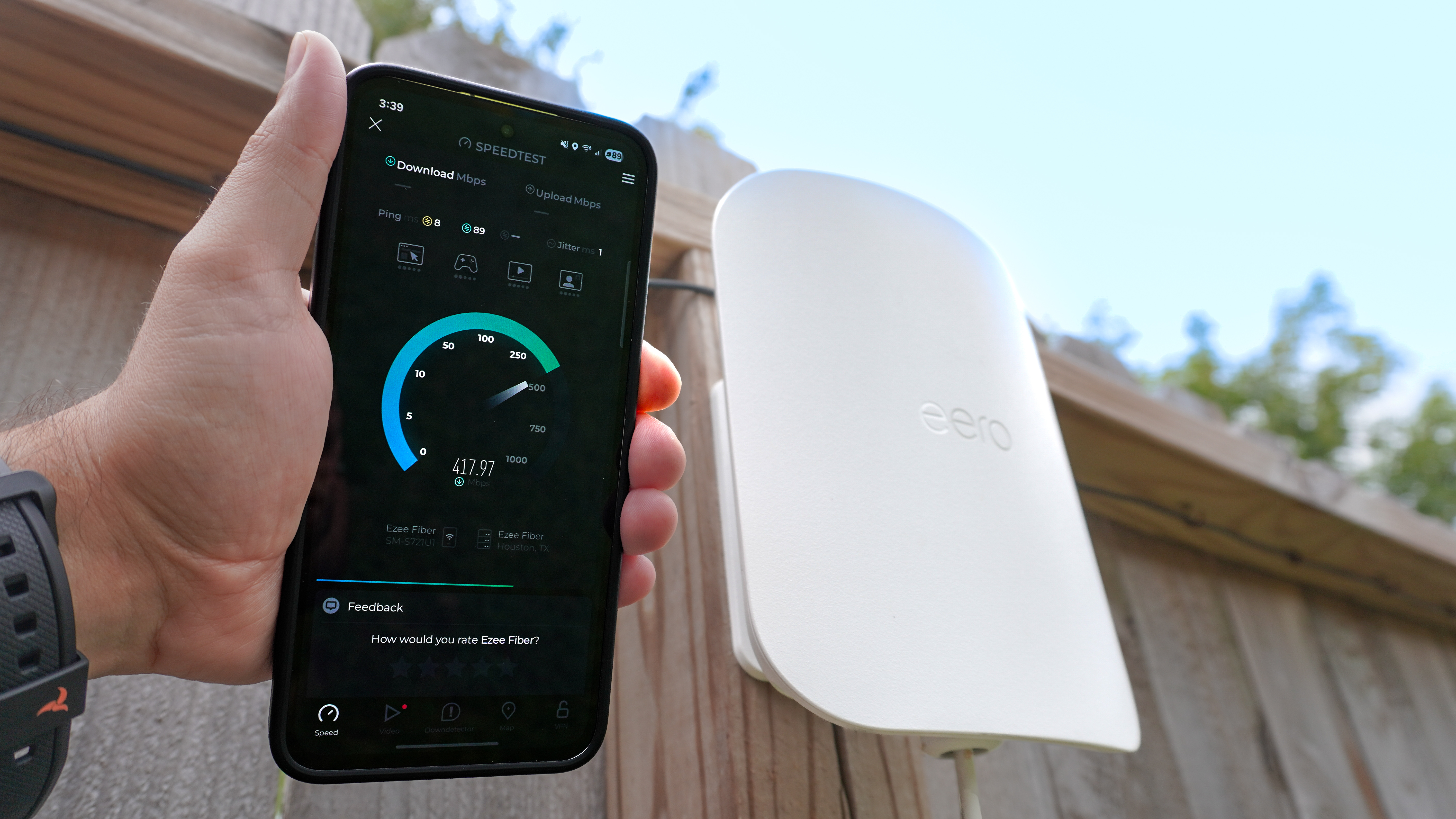 A person holding a phone running an internet speed test in hand with an eero Outdoor 7 mesh extender mounted on a fence in the background