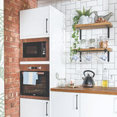 Exposed brick kitchen with white worktops. There is a white built in oven and mircrowave, black kettle and plenty of house plants.