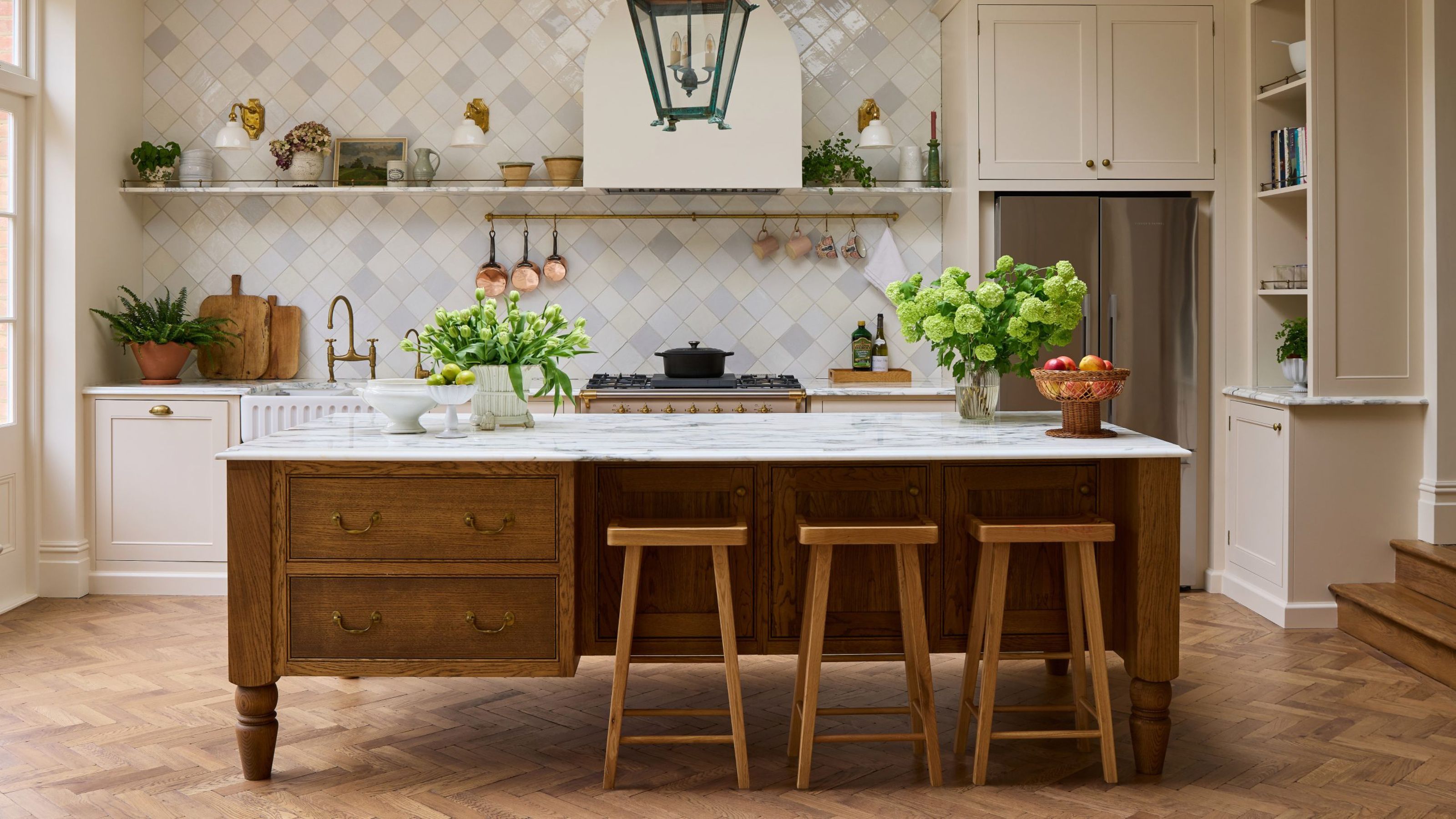 Bright, elegant kitchen featuring a large wooden island with marble top, wooden stools, fresh green flowers, cream cabinets, and a diamond-tiled backsplash on herringbone floors