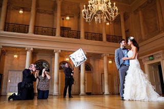 Bride and groom embrace inside grand hall with team of photographers around them