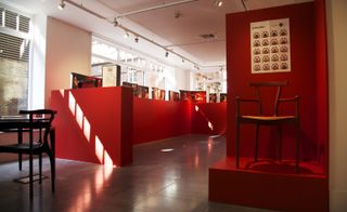 Display room, slate grey floor, white wall and ceiling with spotlights, windows letting in light left side and far wall, red wall surround display with female erotic art along the top, wooden stool on a red viewing platform with artwork above on a red wall front of shot, black table and chair front left