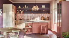 Here is a colorful kitchen island with a dusky pink kitchen island, white countertops, a brown flower pendant light above it, and dark purple walls with a pink vertically tiled splashback