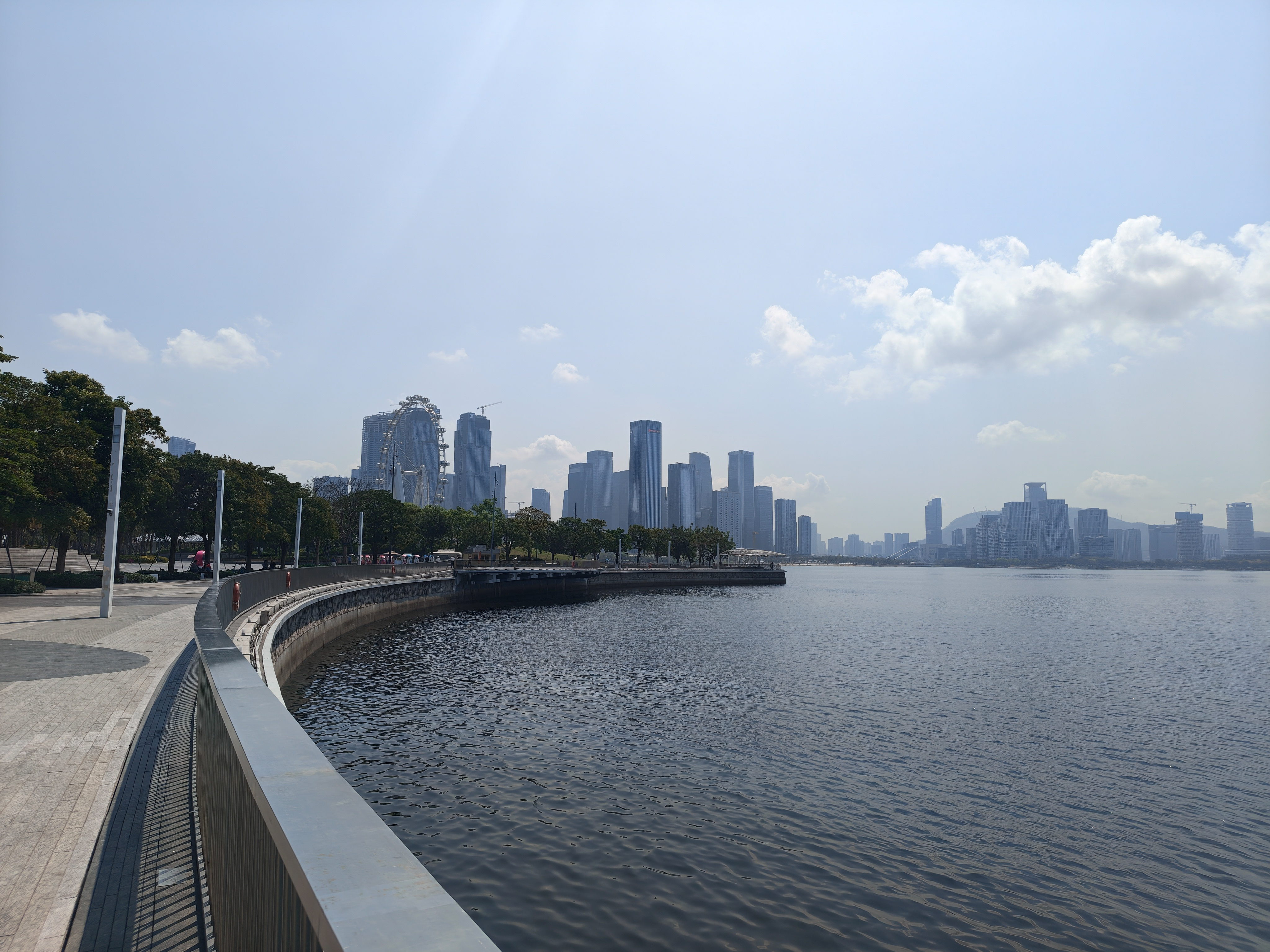 A wide-angle landscape shot of a curved waterfront promenade. A gray metal railing follows the curve of the walkway in the foreground. Across the dark water, a city skyline features various modern skyscrapers and a large white Ferris wheel. The sky is a pale blue with scattered light clouds.