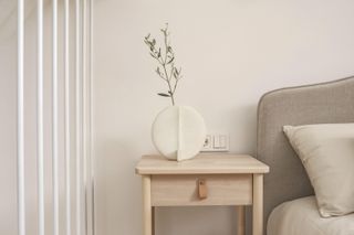 A neutral bedroom with a wooden bedside table decorate with a white ceramic vase a sage green foliage