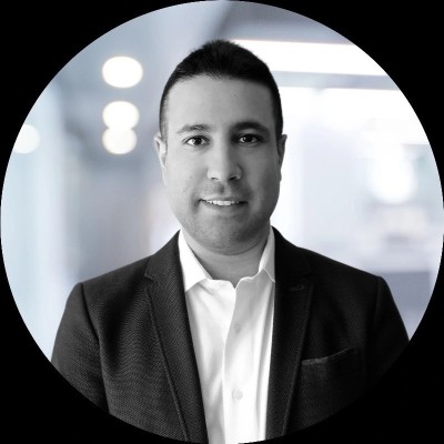 Eli is a man with short hair, pictured in black and white, wearing a collared white shirt and black suit jacket