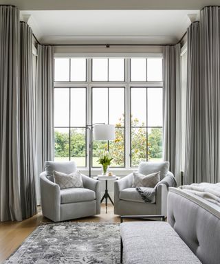 drapes vs blinds, gray bedroom with large bay window, grey floor to ceiling drapes, pair of armchairs in window, rug, bed to the right