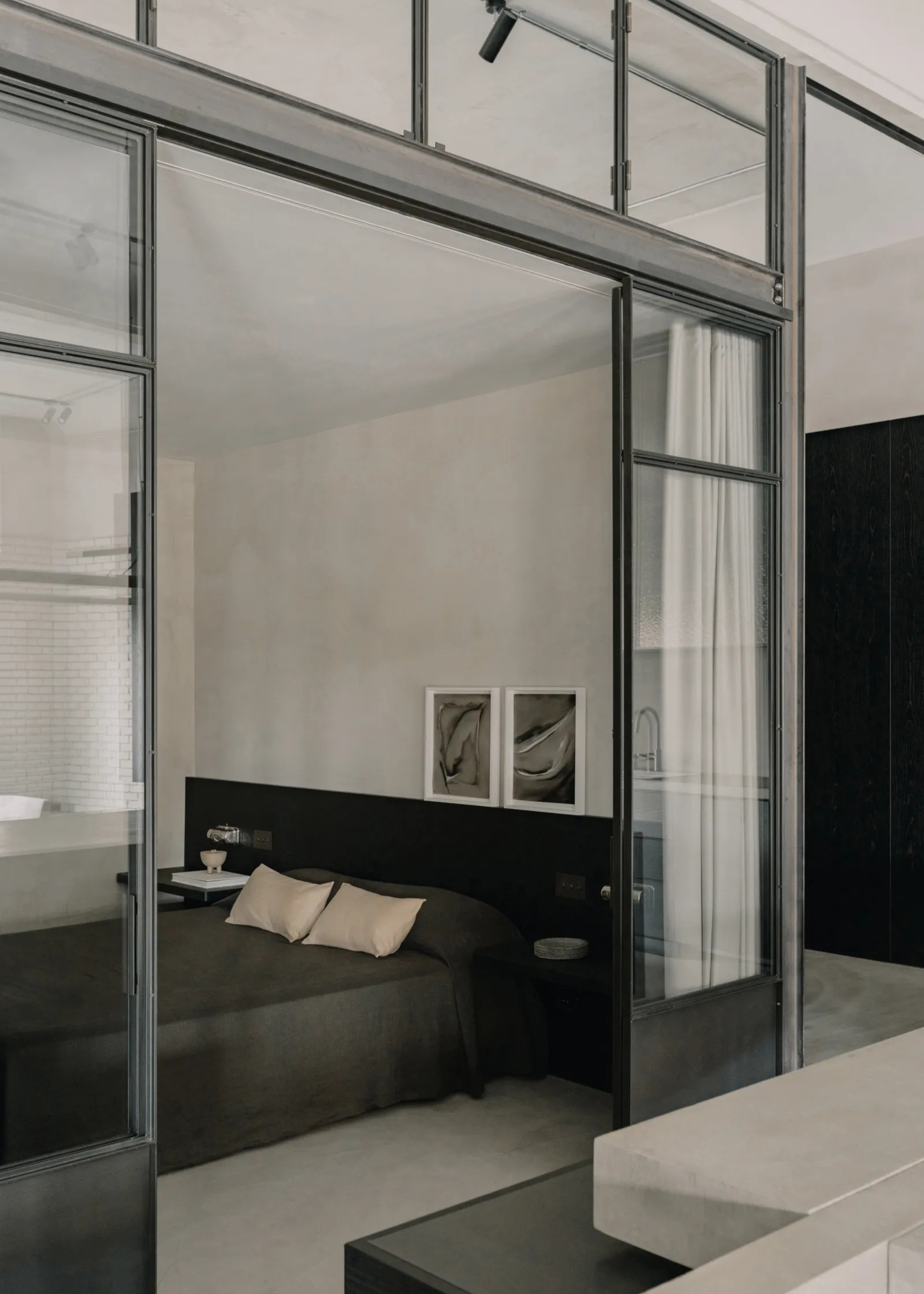 An industrial style bedroom with a low bed in charcoal gray bedding, glass doors with iron framing and a set of framed marbled paintings