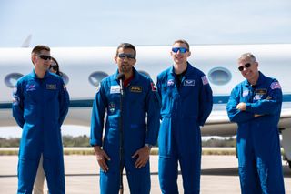 four astronauts in blue spacesuits speak at a press conference at an airport tarmac