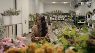 Meghan Markle standing in a florist wearing a camel coat and blue shirt with her arms crossed