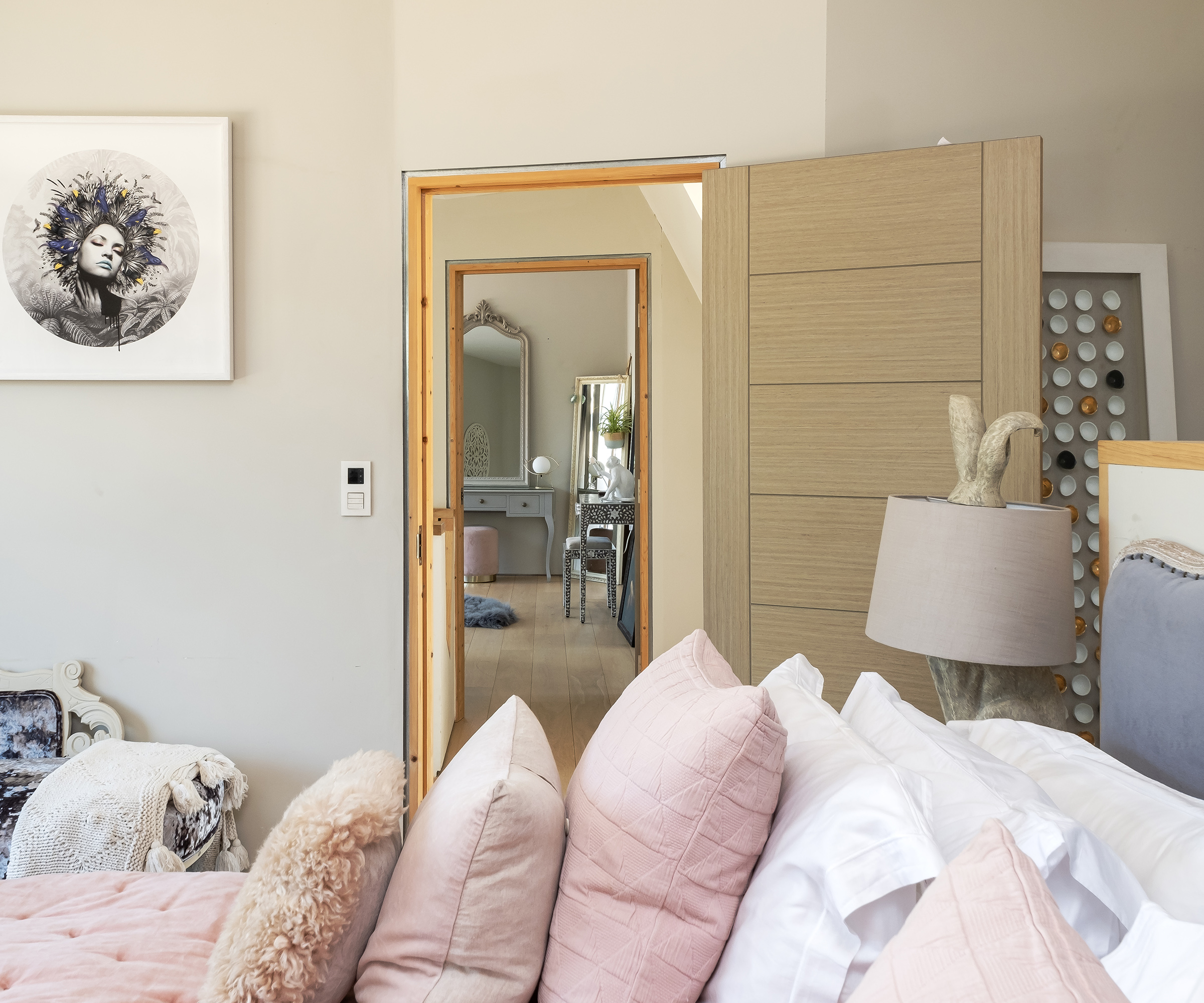 A view through the open door of a bedroom looking to a landing and across the double bed with lots of pastel pink throw cushions