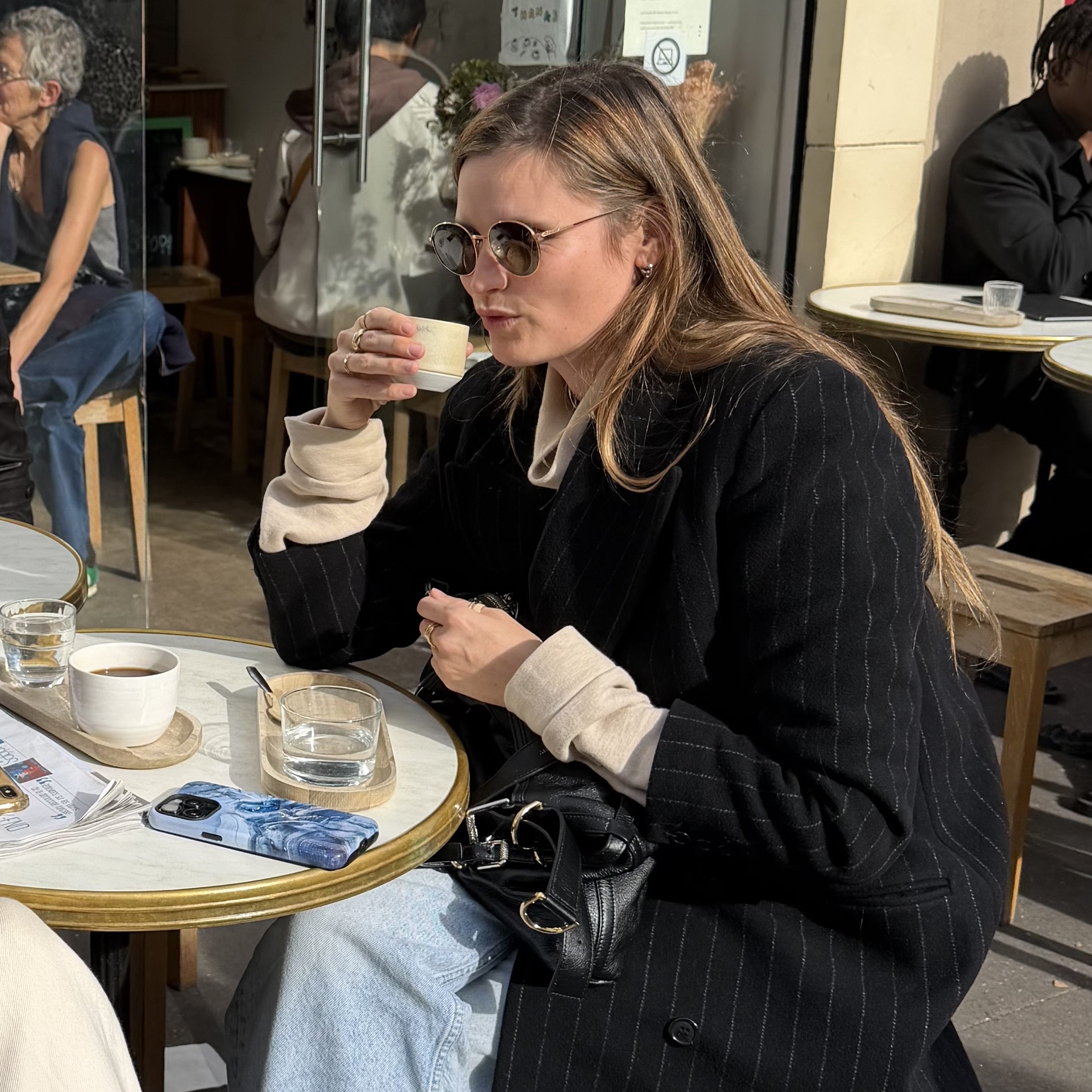 French writer and editor Eugenie Trochu sits at an outdoor cafe in Paris in round sunglasses and a long black coat