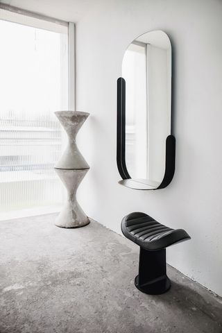 A monochromatic design showroom features brutalist interiors decorated with sculptural furnishings including a ceramic totem, an oblong mirror, and a leather and metal black stool.