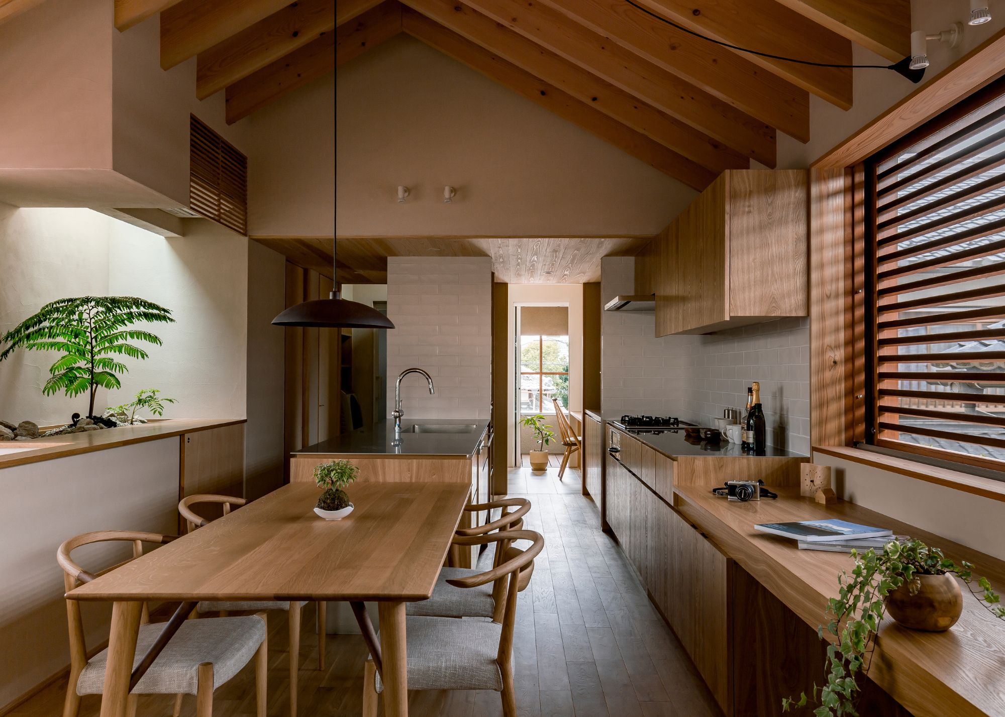 A Japanese kitchen with neutral walls and wooden cabinetry with a dining table that lined up next to the kitchen peninsula with intentional planting that pops against the warm natural materials