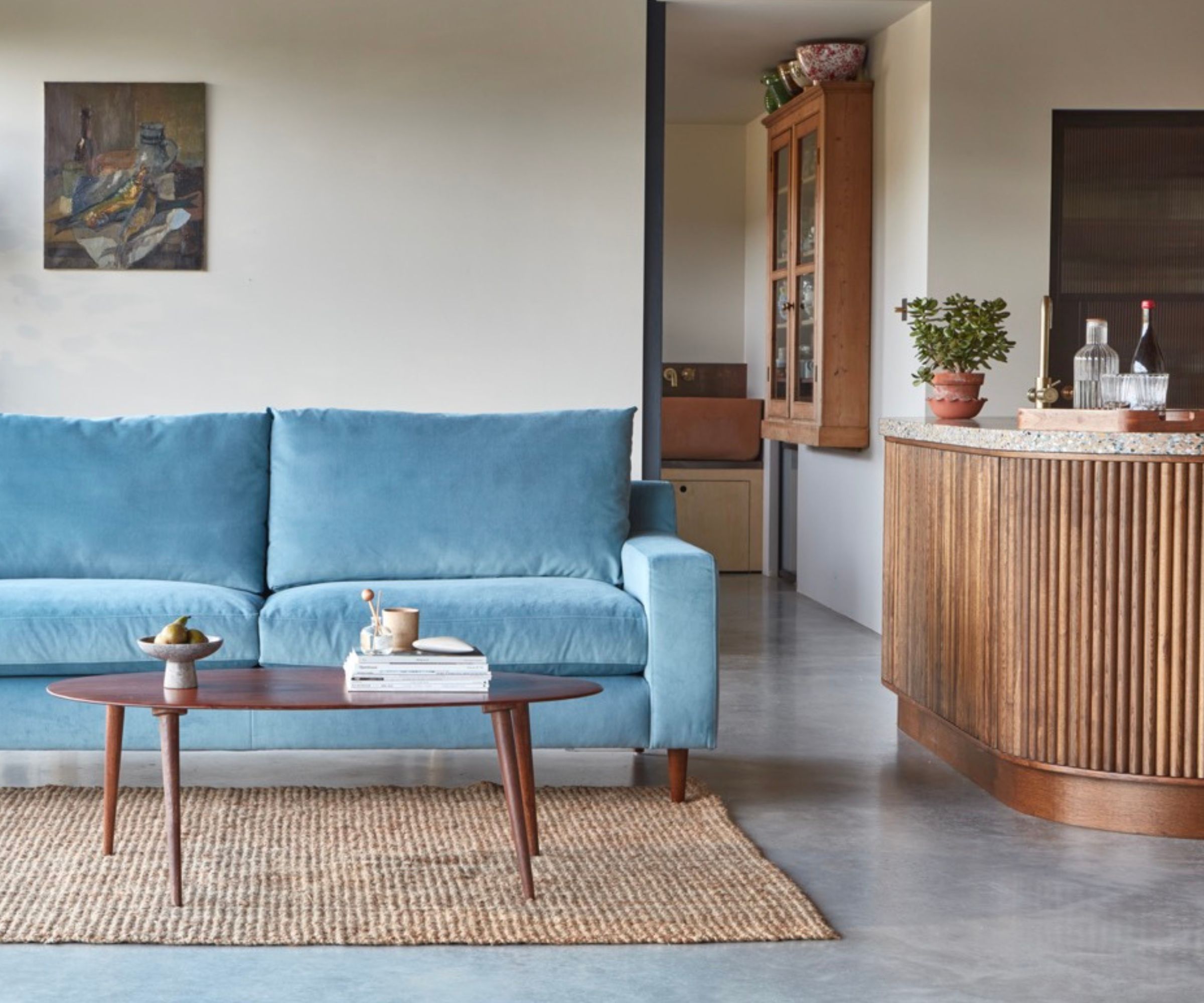 Blue velvet sofa, oval walnut coffee table on jute rug, curved wooden cabinet and glass display case.