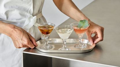 A member of a kitchen brigade captured up close as they handle a silver tray in brushed steel filled with three mini Martinis.