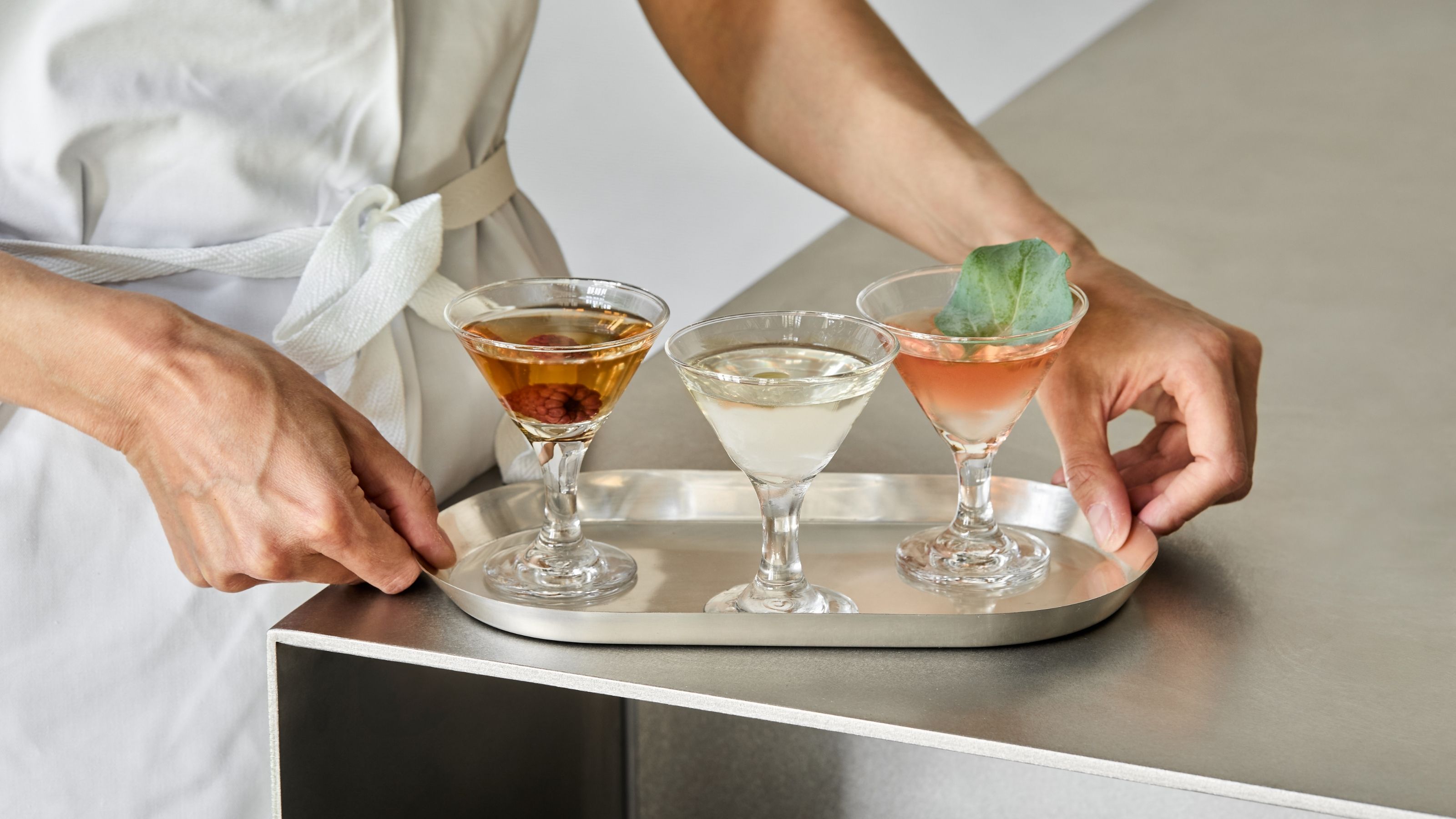 A member of a kitchen brigade captured up close as they handle a silver tray in brushed steel filled with three mini Martinis.