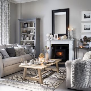 a neutral living room with tones of grey, a fire burning in a woodburner fireplace, light grey sofas with matching cushions and throws and a wooden coffee table on top of a grey patterned rug
