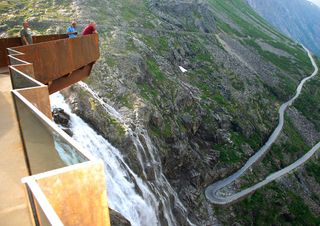 View from a rusted steel viewing platform of the Trollstigen road which winds down the mountainside. There are three people on the viewing platform